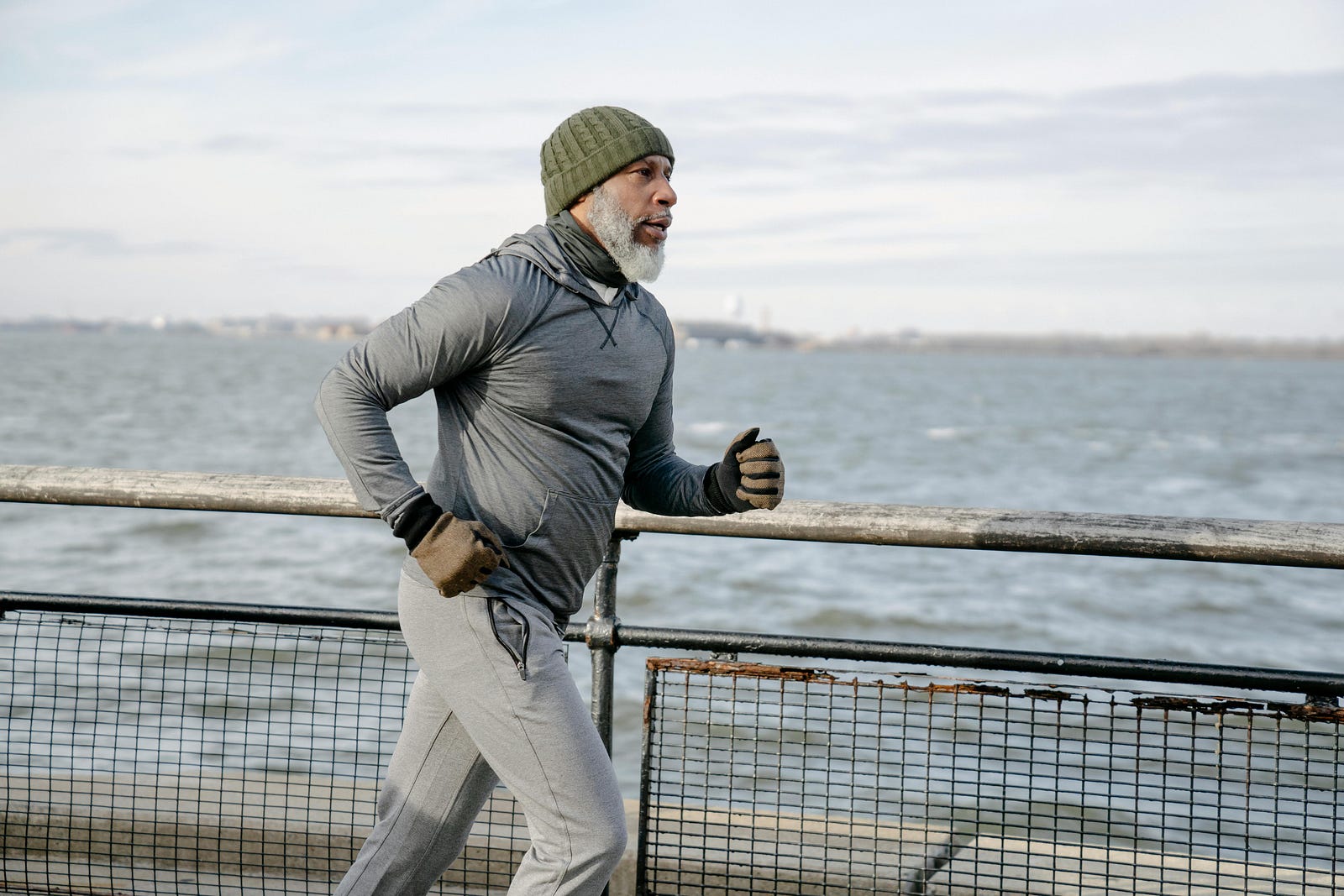 Older man in a beanie, gloves and gray tracksuit running beside a waterfront railing