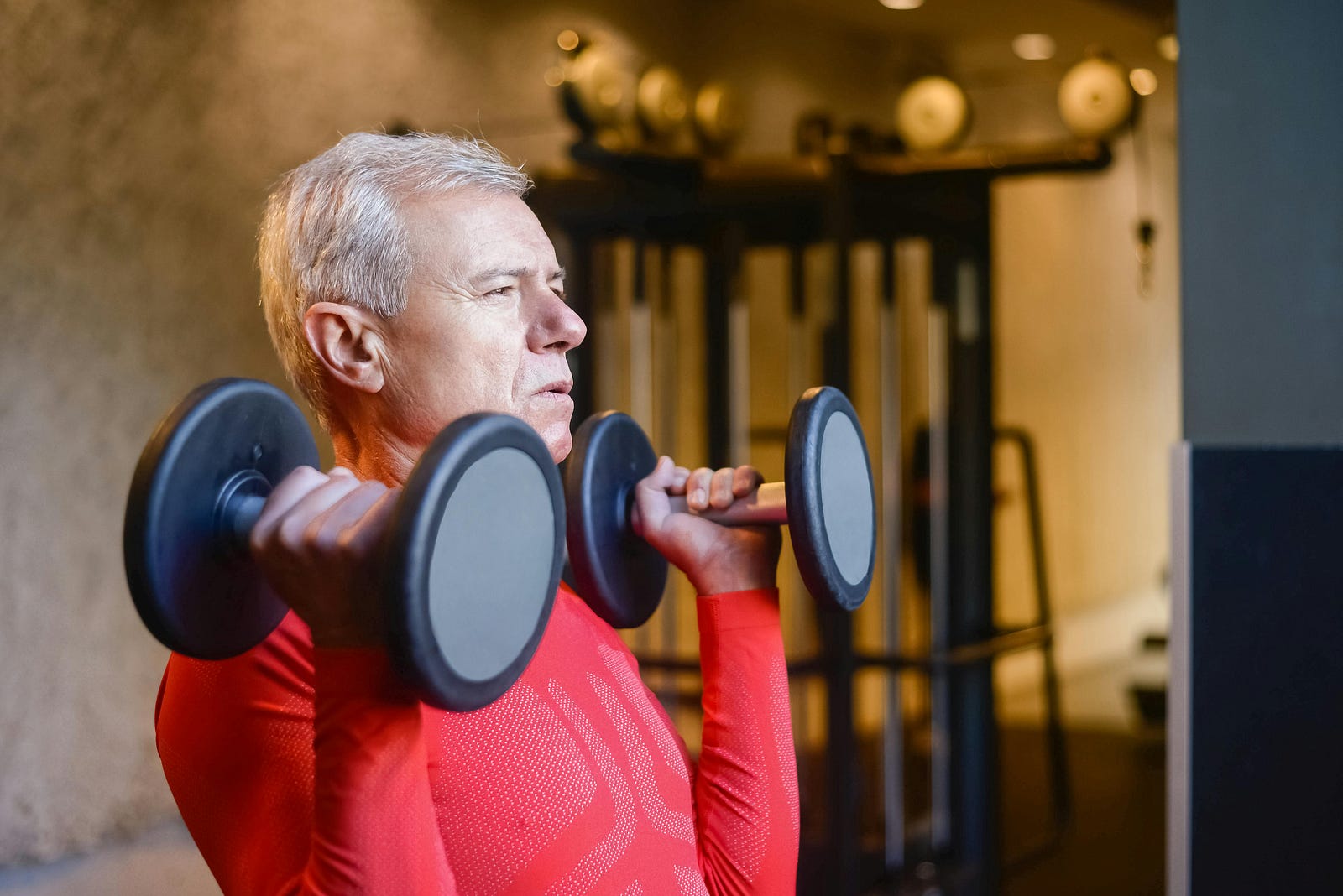 Older man in a gym holding two dumbbells at shoulder height
