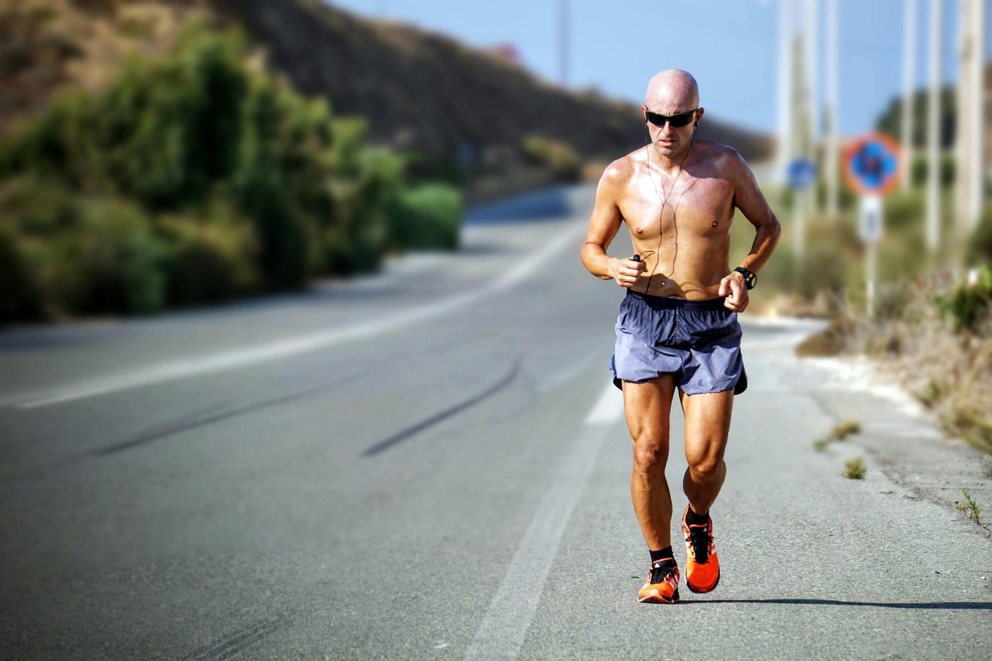 Shirtless runner in sunglasses and earphones running along the shoulder of a sunny road