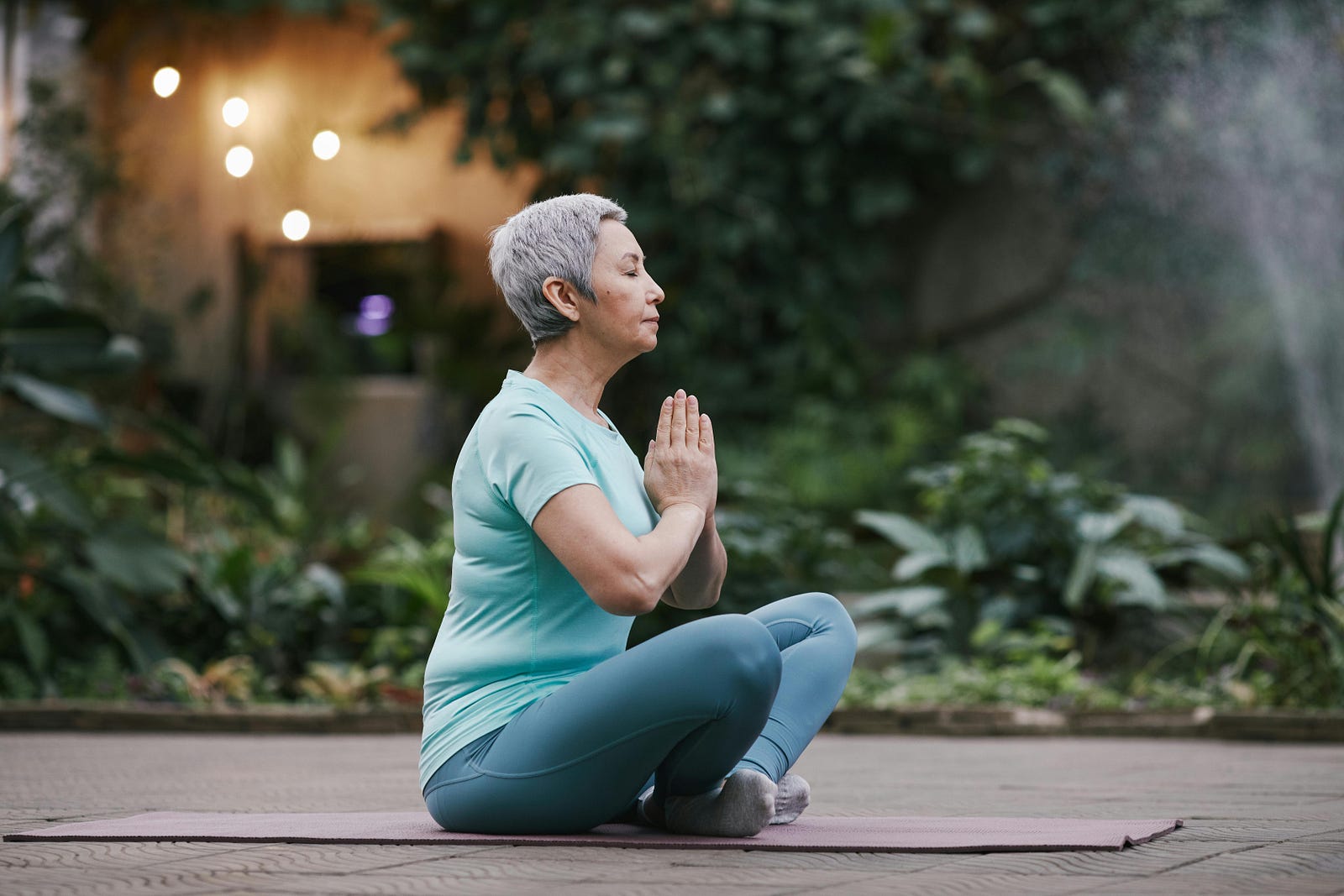 Older woman sitting cross-legged on a yoga mat outdoors, eyes closed and palms pressed together at her chest