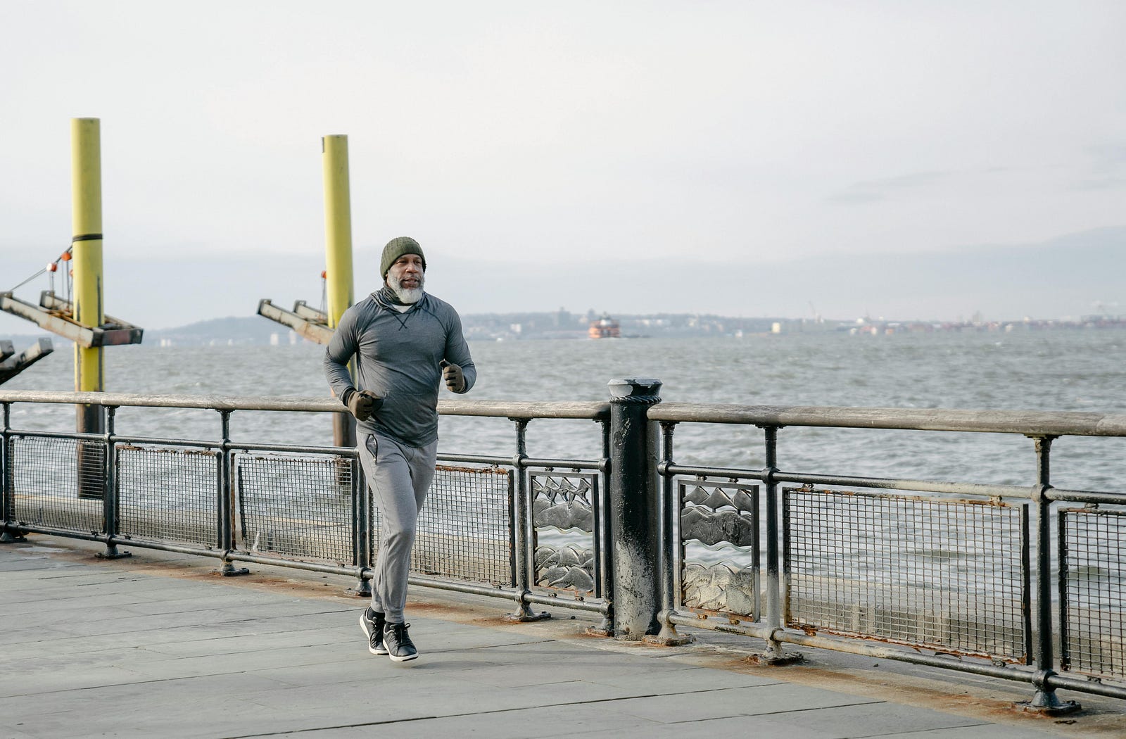 Older man in a beanie jogging along a waterfront promenade on an overcast day