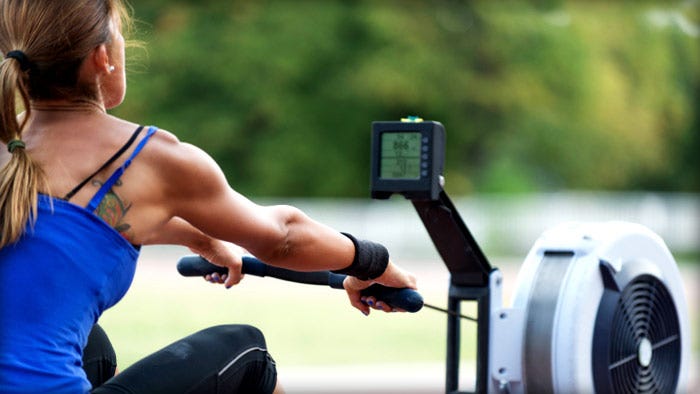 Woman on an indoor rowing machine outdoors, gripping the handle with arms extended toward the monitor