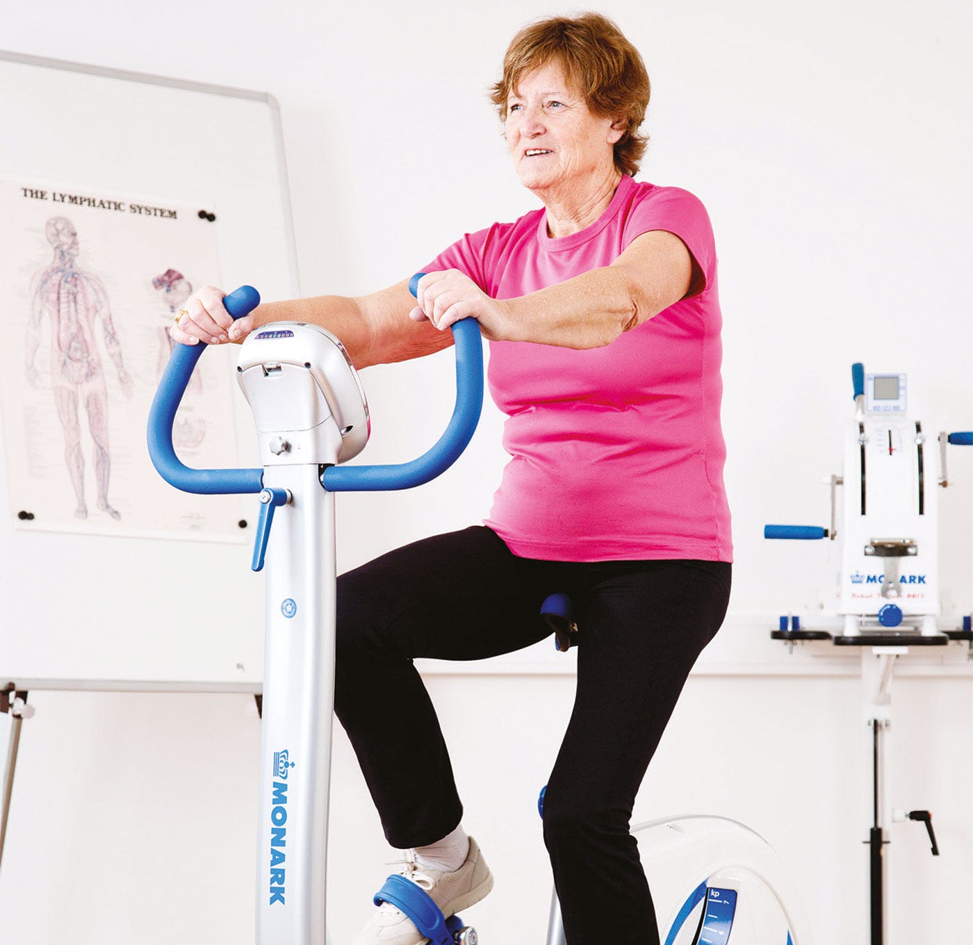 Older woman pedaling an upright exercise bike in a clinic, with an arm-crank ergometer in the background