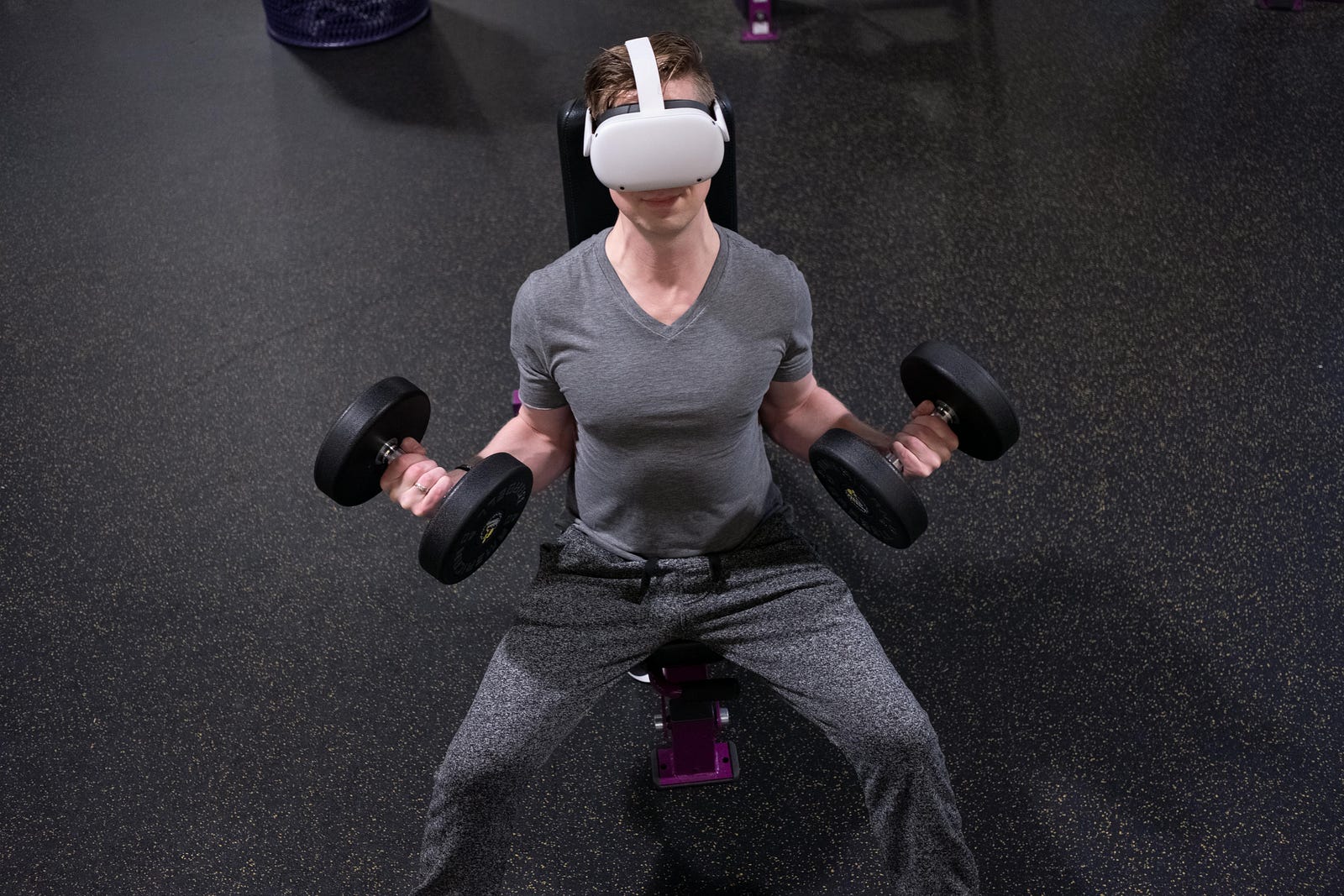 Man wearing a VR headset doing seated dumbbell curls on a bench in a gym