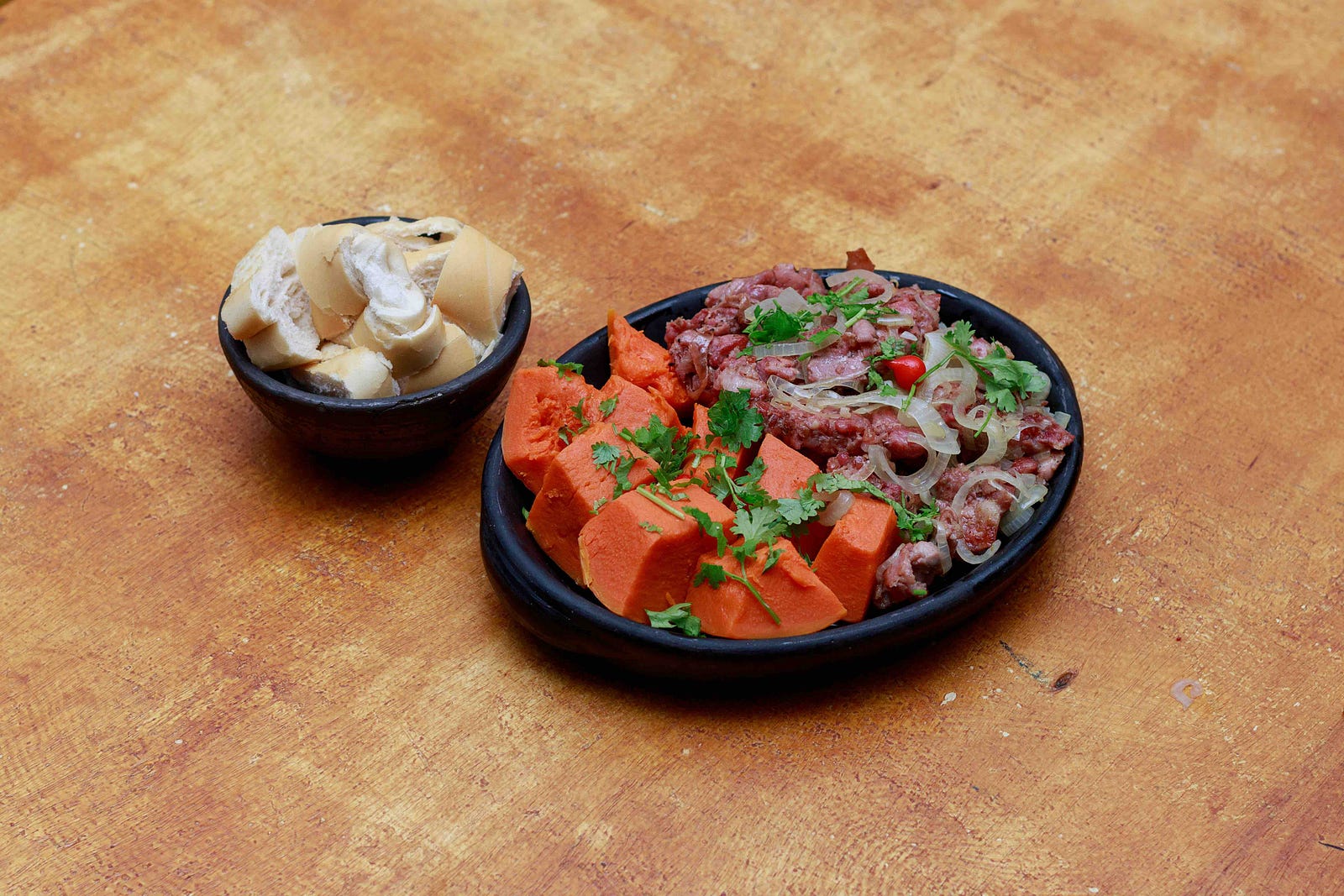 Plate of cooked orange squash or sweet potato with sliced meat, onions and cilantro, beside a bowl of bread