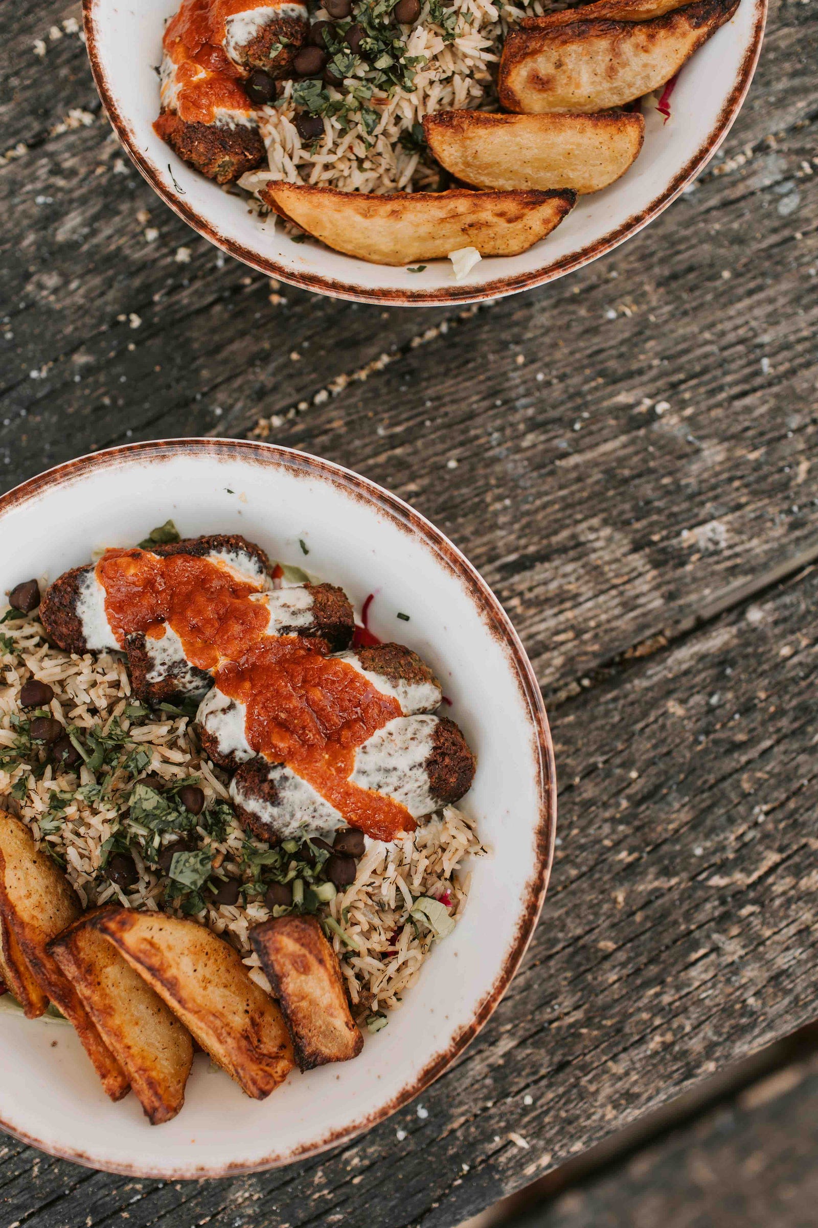 Two bowls of herbed rice with chickpeas, falafel topped with red and white sauces, and roasted potato wedges